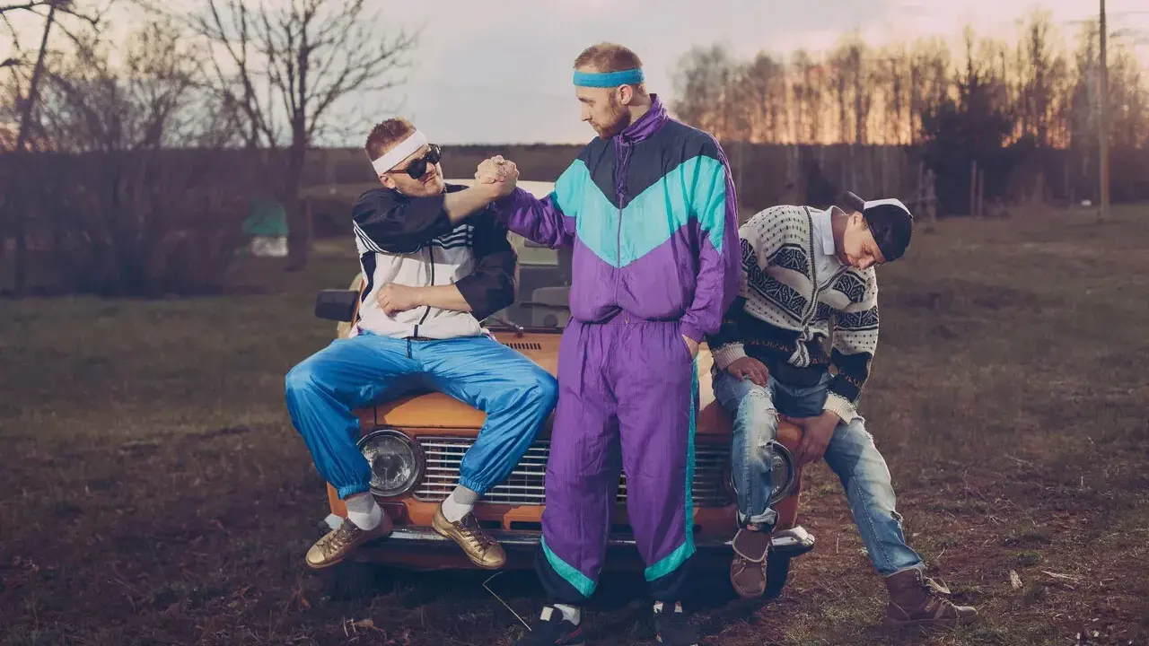 Trzech mężczyzn w dresach i swetrach, styl grunge lata 90., siedzi na masce starego samochodu.