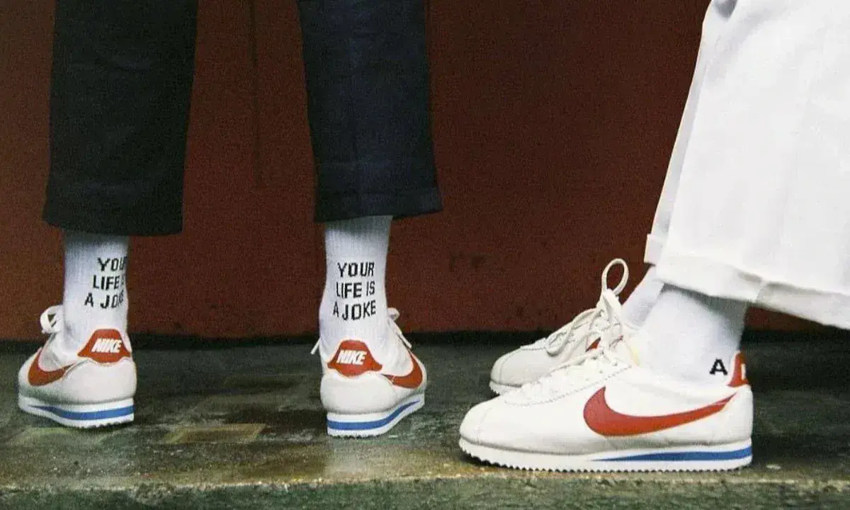 Stare modele butów Nike Cortez w klasycznej odsłonie. Białe z czerwonym logo i niebieskim paskiem. Na skarpetkach napis "YOUR LIFE IS A JOKE".