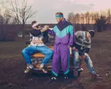 Trzech mężczyzn w dresach i swetrach, styl grunge lata 90., siedzi na masce starego samochodu.