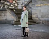 Kobieta w płaszczu w pistacjowym kolorze, z beżową torebką i ozdobnymi butami, stoi przed budynkiem Deutsche Staatsoper.