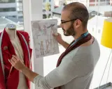 Mężczyzna z wąsami i okularami, wpatrzony w szkic sukienek, przygotowuje się do stworzenia kolekcji jak najsłynniejsi projektanci mody.