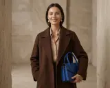 Elegancka kobieta w brązowym płaszczu i beżowej bluzce trzyma niebieską torebkę. Idealny przykład, jaki kolor pasuje do brązu ubrania.