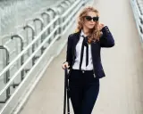 Eleganckie stylizacje z czarnymi spodniami: kobieta w ciemnej kurtce, białej koszuli z kokardą i okularach przeciwsłonecznych idzie po moście.