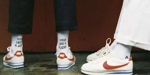 Stare modele butów Nike Cortez w klasycznej odsłonie. Białe z czerwonym logo i niebieskim paskiem. Na skarpetkach napis "YOUR LIFE IS A JOKE".