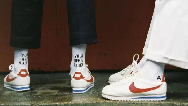 Stare modele butów Nike Cortez w klasycznej odsłonie. Białe z czerwonym logo i niebieskim paskiem. Na skarpetkach napis "YOUR LIFE IS A JOKE".
