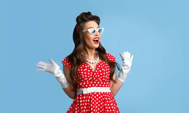 Kobieta w czerwonej sukience w białe grochy, białych rękawiczkach i okularach w stylu pin-up girl stylizacja, z uniesionymi rękami.