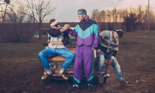 Trzech mężczyzn w dresach i swetrach, styl grunge lata 90., siedzi na masce starego samochodu.