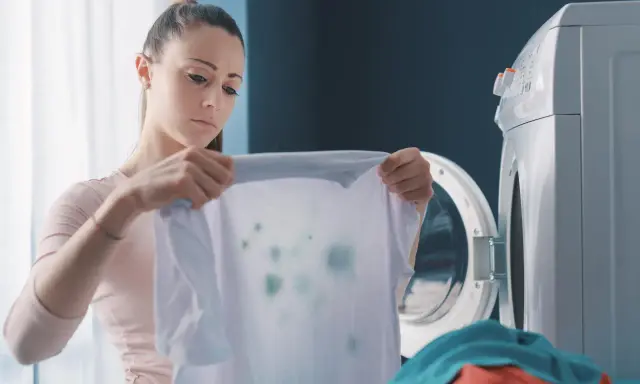 Zmartwiona kobieta trzyma białą koszulkę z zielonymi plamami, która wygląda jak zafarbowane ubrania. Obok stoi otwarta pralka.