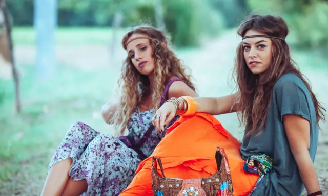 Dwie dziewczyny w stylu lat 70. moda impreza, z opaskami na głowach i kwiecistymi sukienkami, siedzą na trawie.