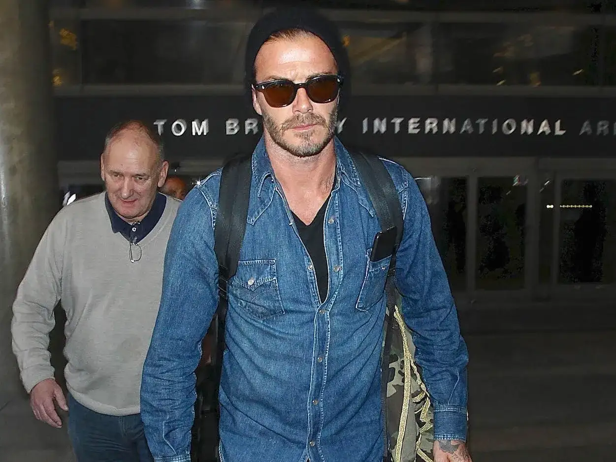 David Beckham w jeansowej koszuli i czapce, inspiracja dla męskich stylizacji. Obok mężczyzna w szarym swetrze.