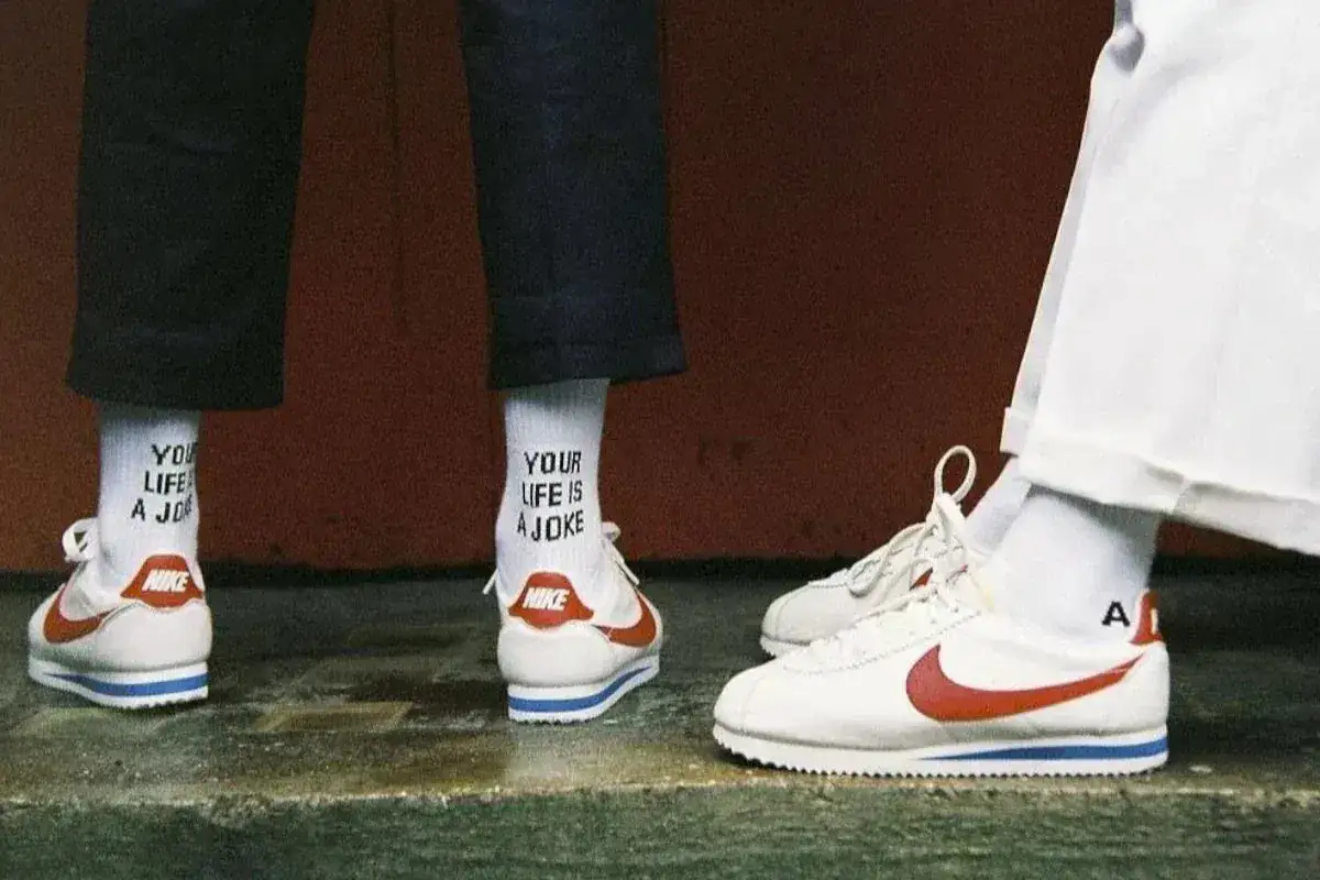 Stare modele butów Nike Cortez w klasycznej odsłonie. Białe z czerwonym logo i niebieskim paskiem. Na skarpetkach napis 