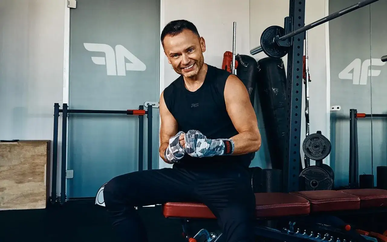 Igor Klaja 4f w sportowym stroju, z rękami owiniętymi bandażami, siedzi na ławce w siłowni.