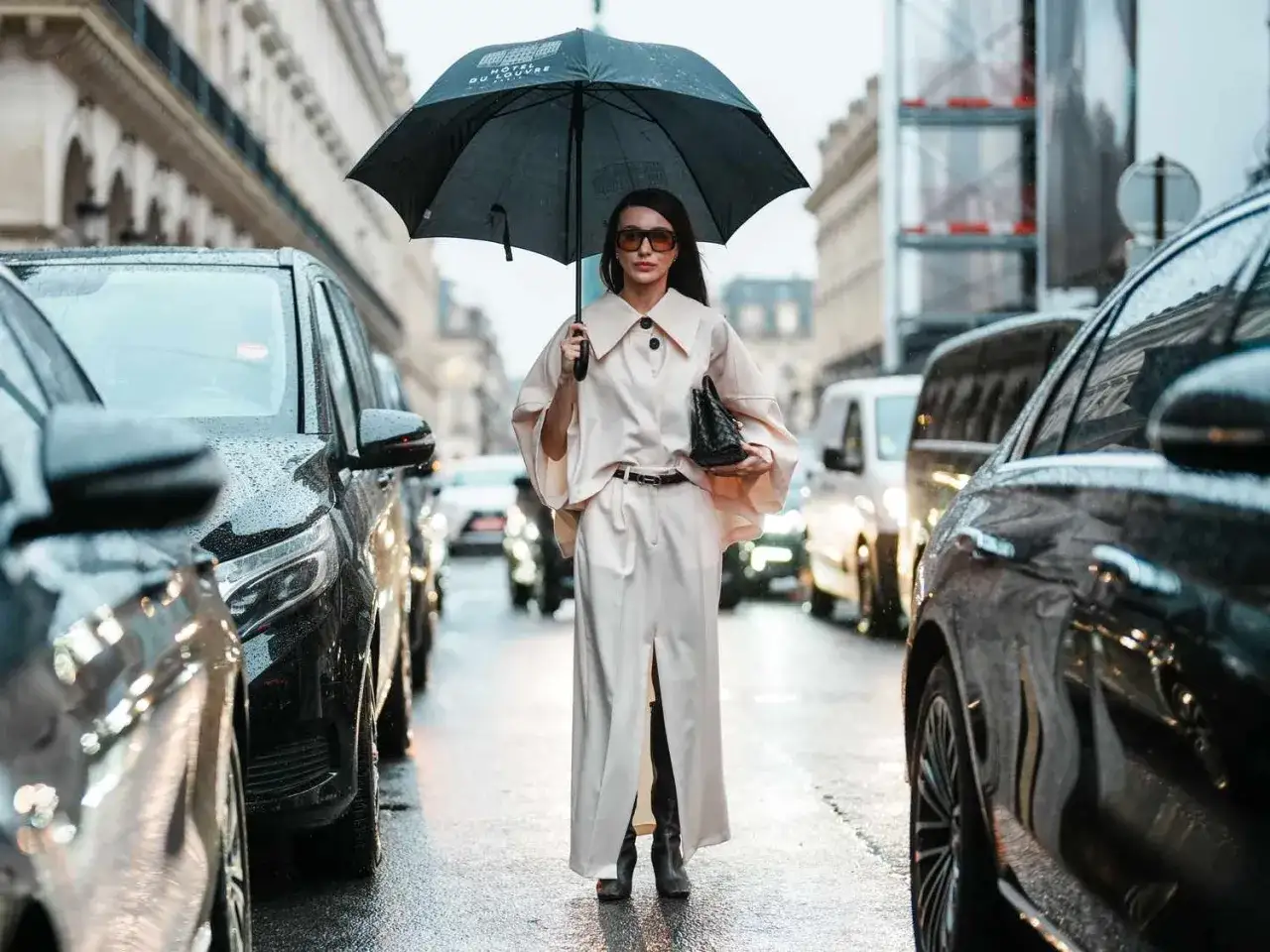 Elegancka kobieta w długim płaszczu i spodniach, z parasolką, prezentuje stylowe stylizacje na deszczowe dni, otoczona samochodami na mokrej ulicy.