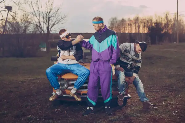 Trzech mężczyzn w dresach i swetrach, styl grunge lata 90., siedzi na masce starego samochodu.