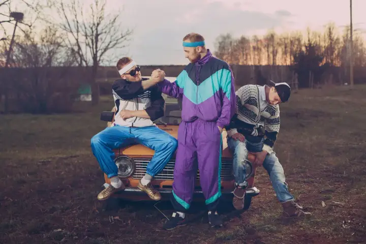 Trzech mężczyzn w dresach i swetrach, styl grunge lata 90., siedzi na masce starego samochodu.